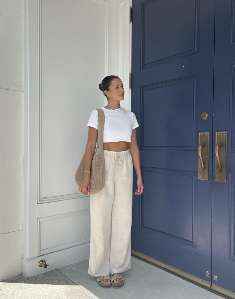 A woman stands poised before a grand blue door with brass fixtures, surrounded by a white wall. She dons a cropped white t-shirt and high-waisted light beige linen pants, creating a relaxed yet polished look. She complements her outfit with simple strappy sandals and a woven shoulder bag. Her hair is sleekly pulled back into a low bun, and she gazes off to the side with a calm and contemplative expression.